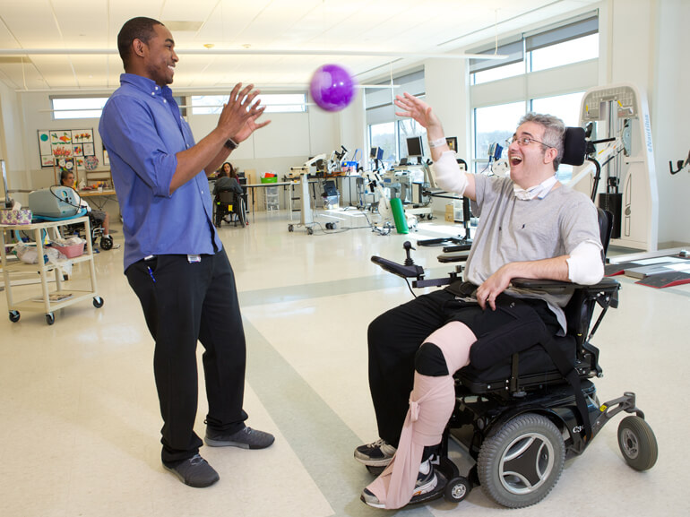 Patient in wheelchair tossing small purple ball to a therapist