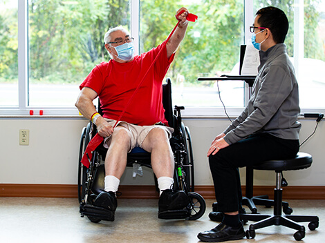 White-haired man sitting in a wheelchair using a red resistance band to do arm exercises.