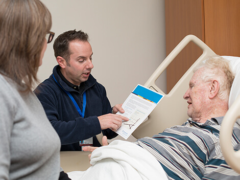 Male therapist holding an admission form while talking to a senior-age man in hospital bed.