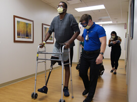 Man with prosthetic leg using rolling walker in hospital hallway.