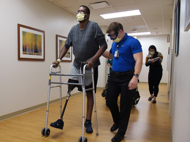 Man wearing leg prosthetic using a rolling walker in hospital hallway.