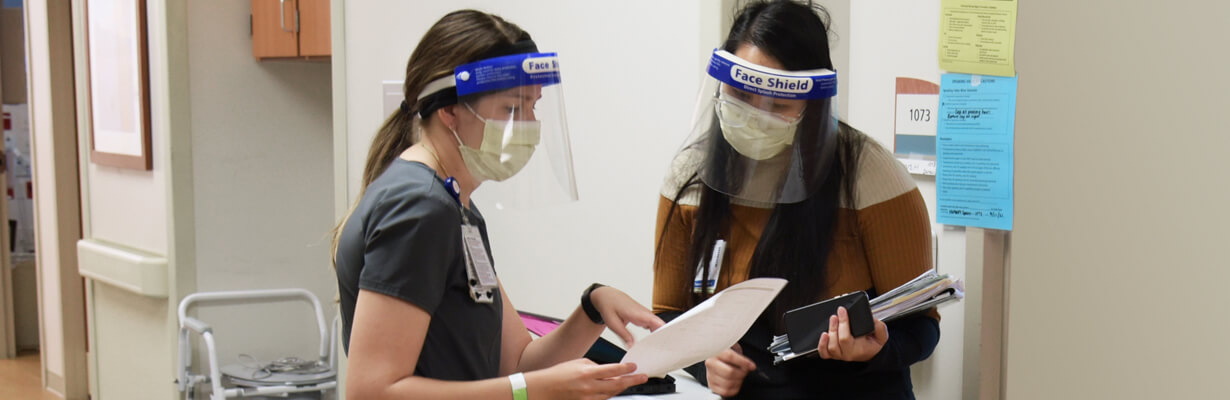 Two female nurses wearing PPE masks with plastic face shields discussing a patient file.