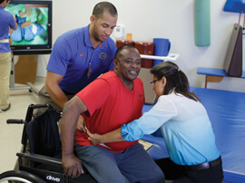 A male and female therapist helping a male patient stand up from his wheelchair.