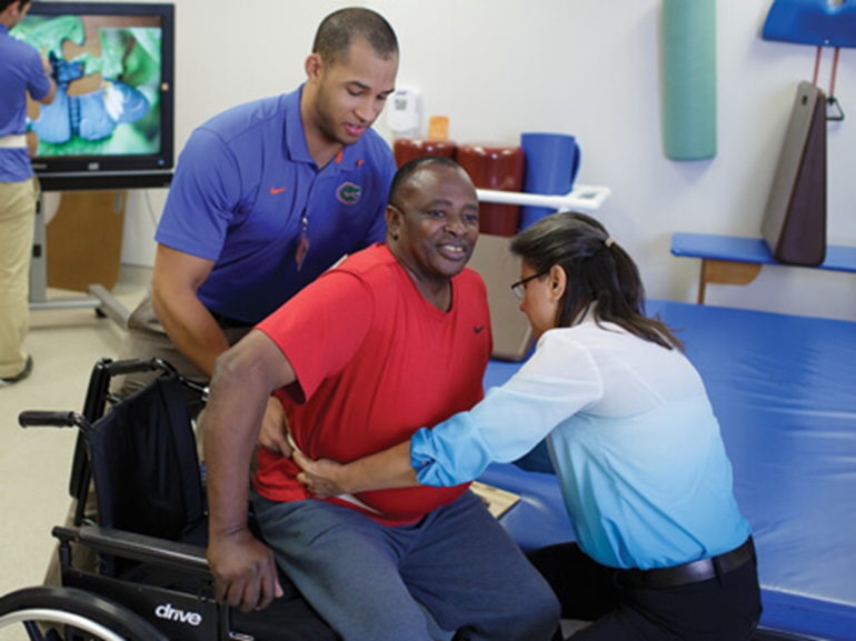 A male and female therapist help a male patient get out of his wheelchair.