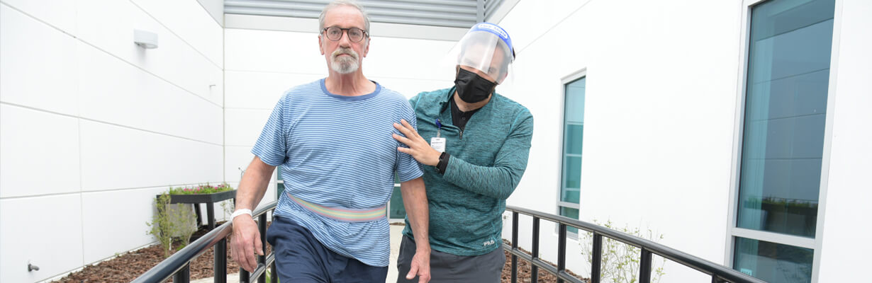 Female therapist wearing PPE face mask helping a male patient walk outside.