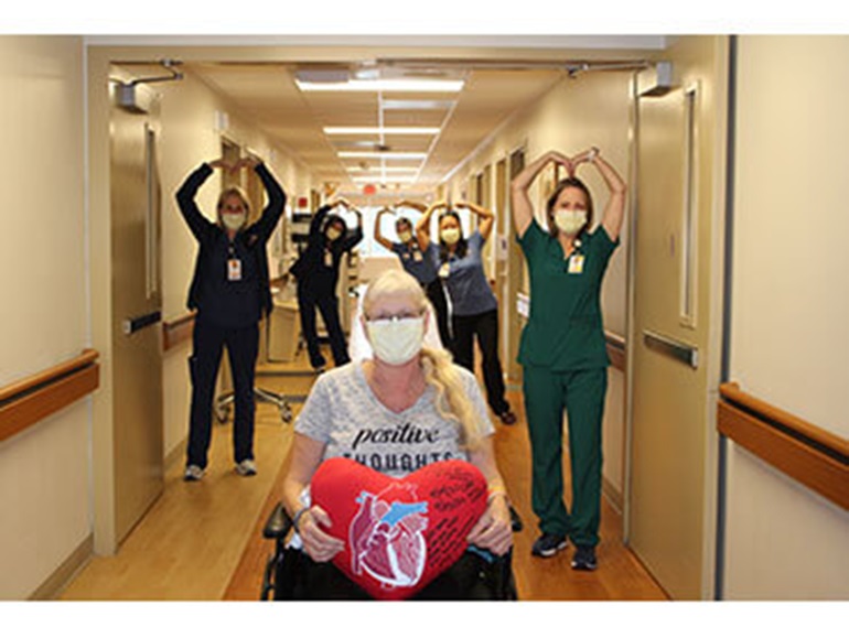 Debra sitting in a wheelchair holding a heart-shaped pillow with nurses in background make heart shapes with their arms.