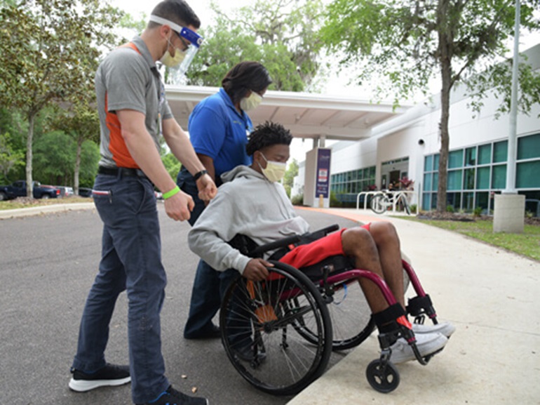 Two therapists helping young male patient get his wheelchair over a curb and onto the sidewalk outside the hospital.